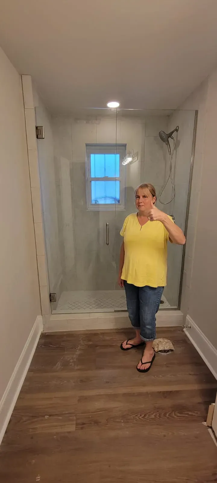 Woman in yellow top and jeans gives thumbs up in a bathroom with a glass shower.