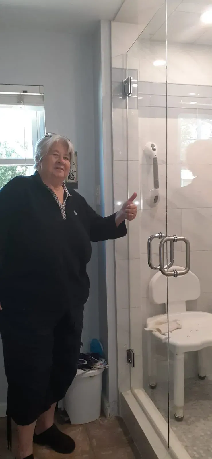 Woman pointing to a grab bar in a walk-in shower. Bathroom with a shower seat and glass door.