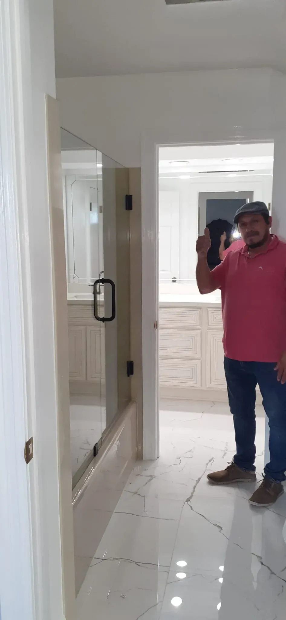 A man in a pink shirt and jeans stands in a hallway. The hallway leads to a bathroom.