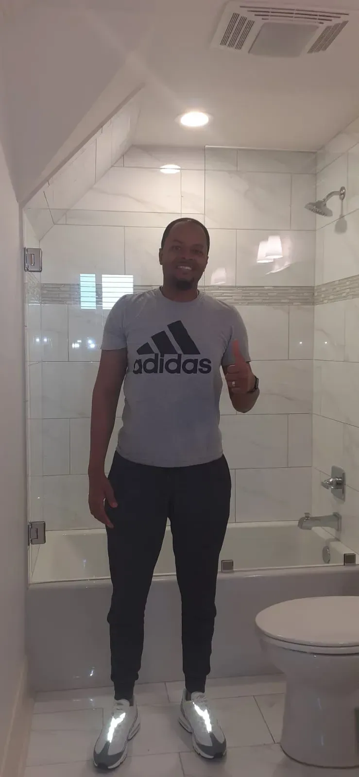 A person standing in a bathroom, wearing an Adidas shirt and black pants, smiling and pointing.