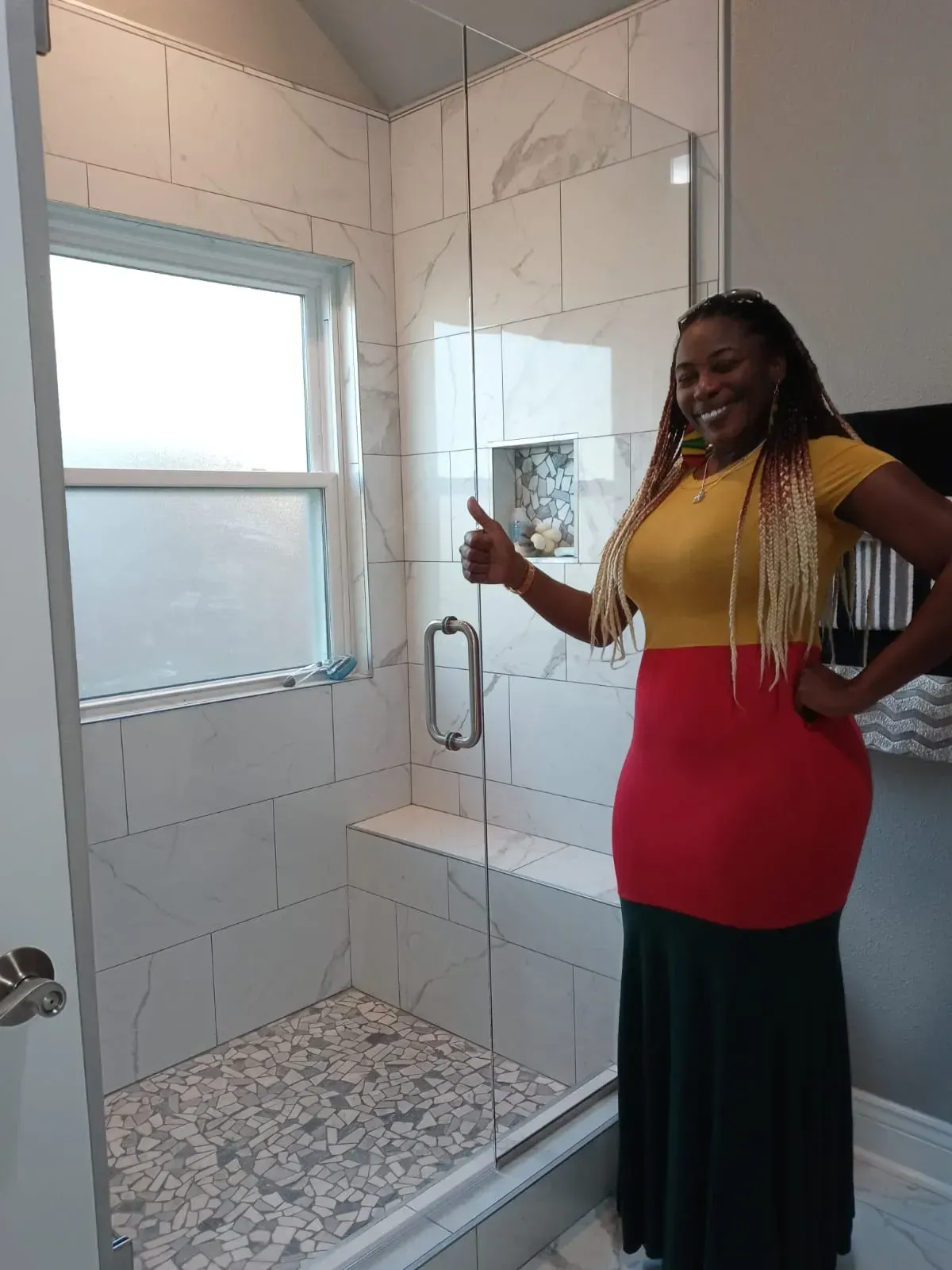 Woman gives thumbs up in a newly tiled shower, with glass door and window. She's wearing a yellow/red/green dress.