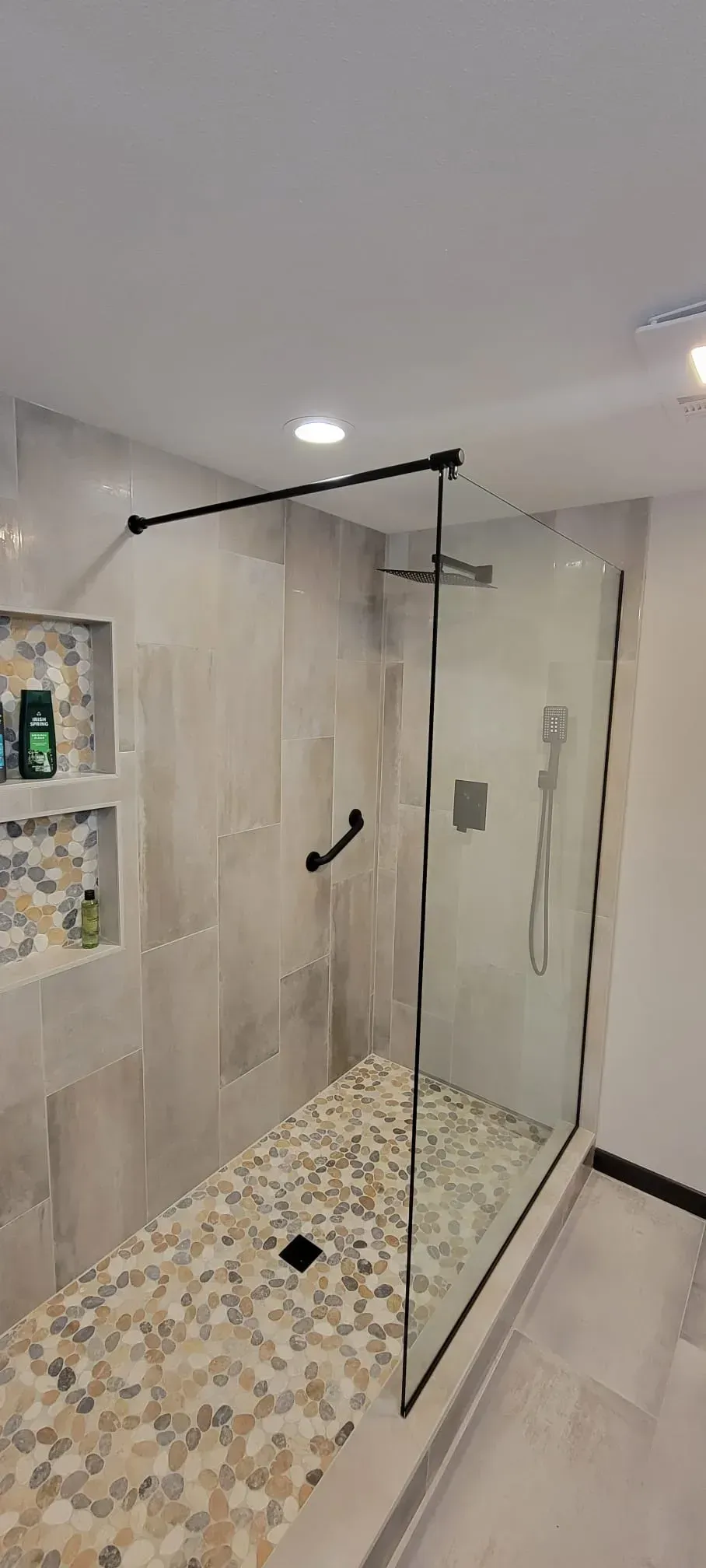 A modern shower with glass walls, black hardware, and pebble flooring. Light gray tile walls.