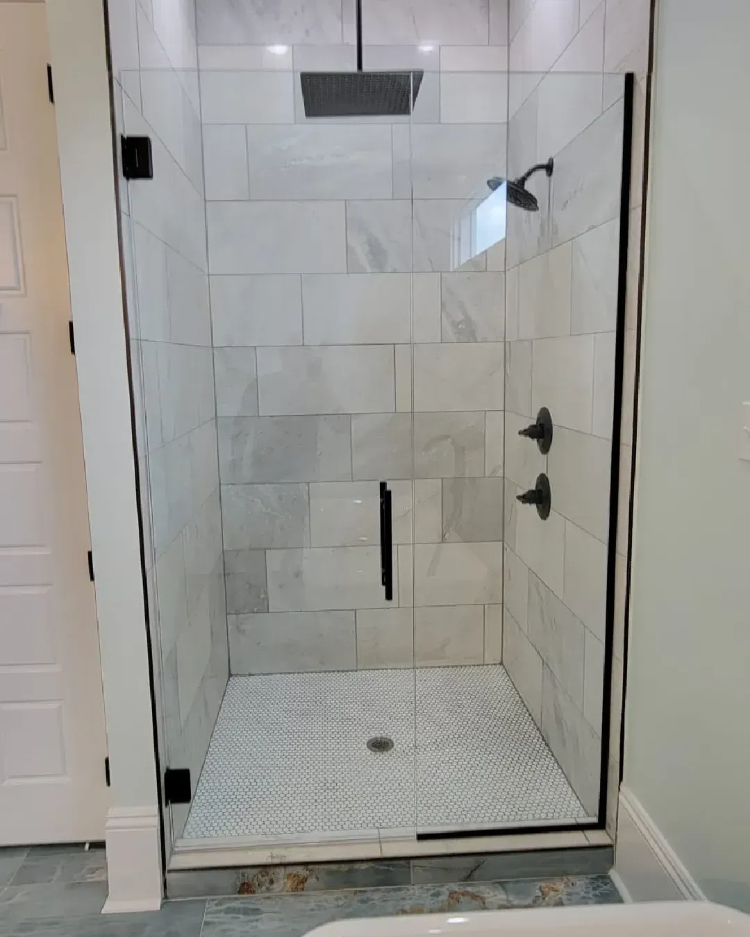 Glass-enclosed shower with white marble-like tiles and mosaic floor. Black fixtures; large overhead showerhead.