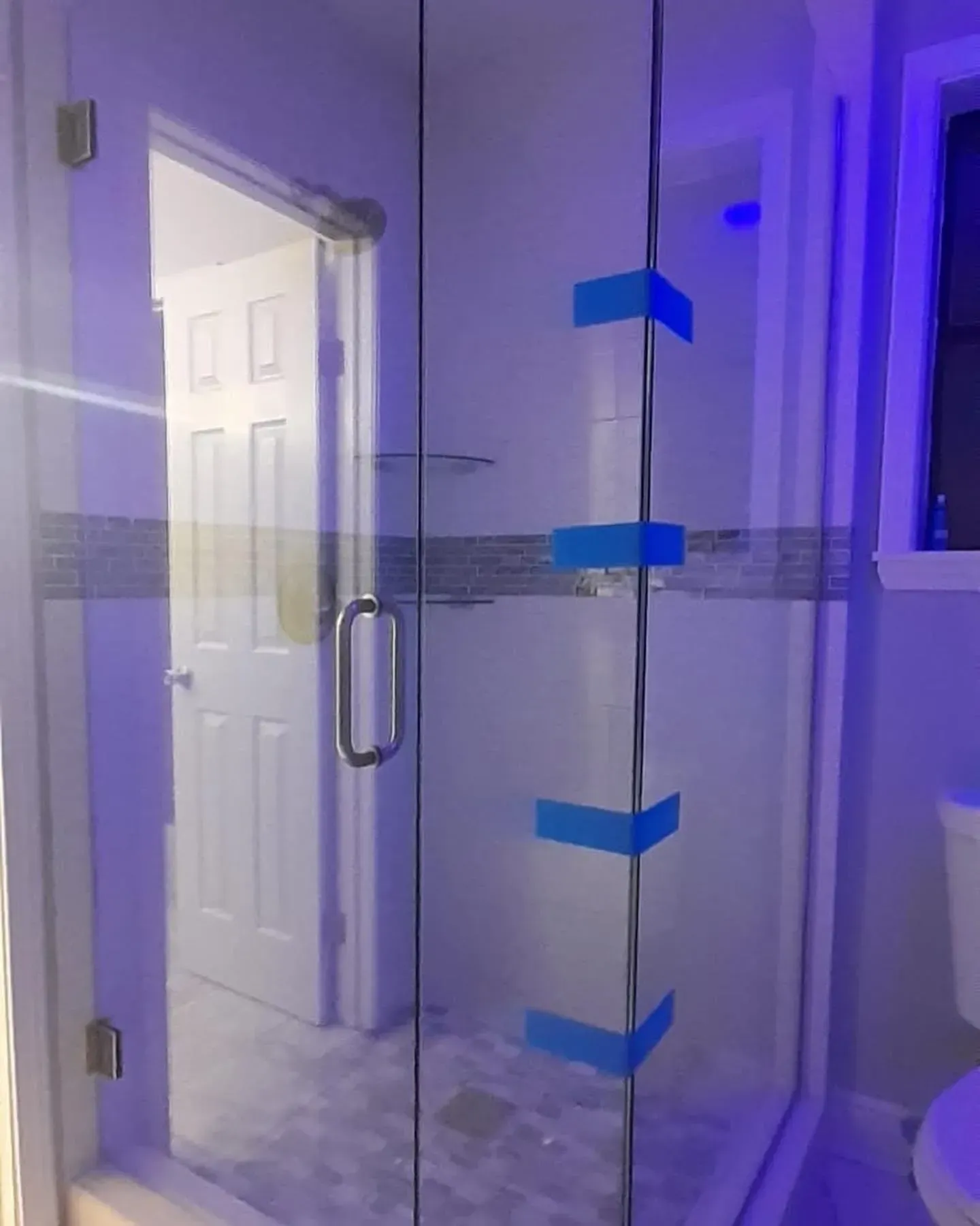 Glass shower door with blue tape, looking into a bathroom with a door and a toilet.