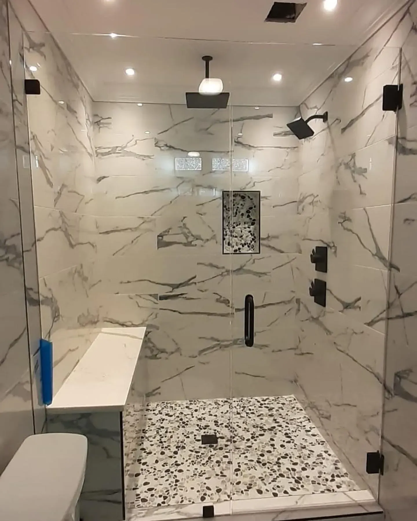 Modern shower with marble-like tile walls and pebble floor. Glass doors and black fixtures.
