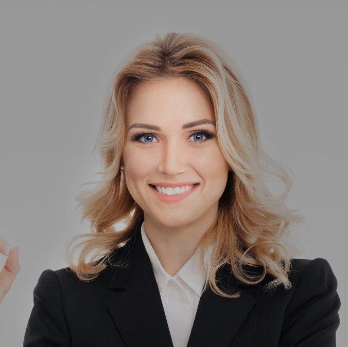 Blonde woman in black blazer smiling at the camera, wearing white shirt. Gray background.