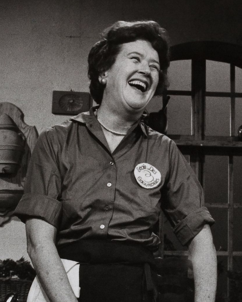 Julia Child laughing, wearing a collared shirt and apron, in a kitchen setting.