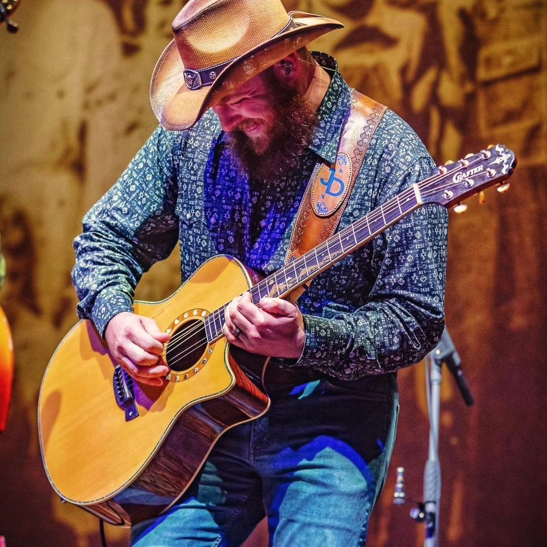Man in cowboy hat playing an acoustic guitar on stage, with a microphone behind him.