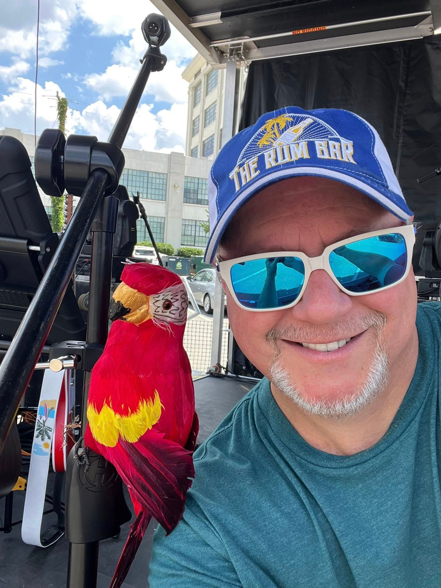 Man in a hat and sunglasses smiles with a red parrot on his shoulder, outdoors near equipment.