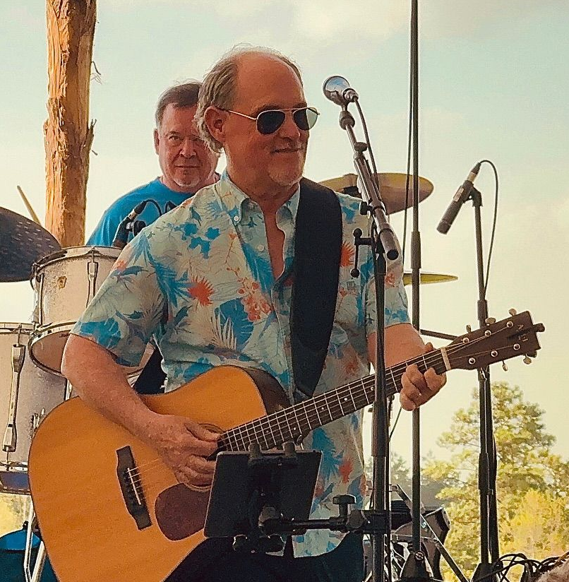 Man playing acoustic guitar on stage, wearing sunglasses, floral shirt; drummer in background.