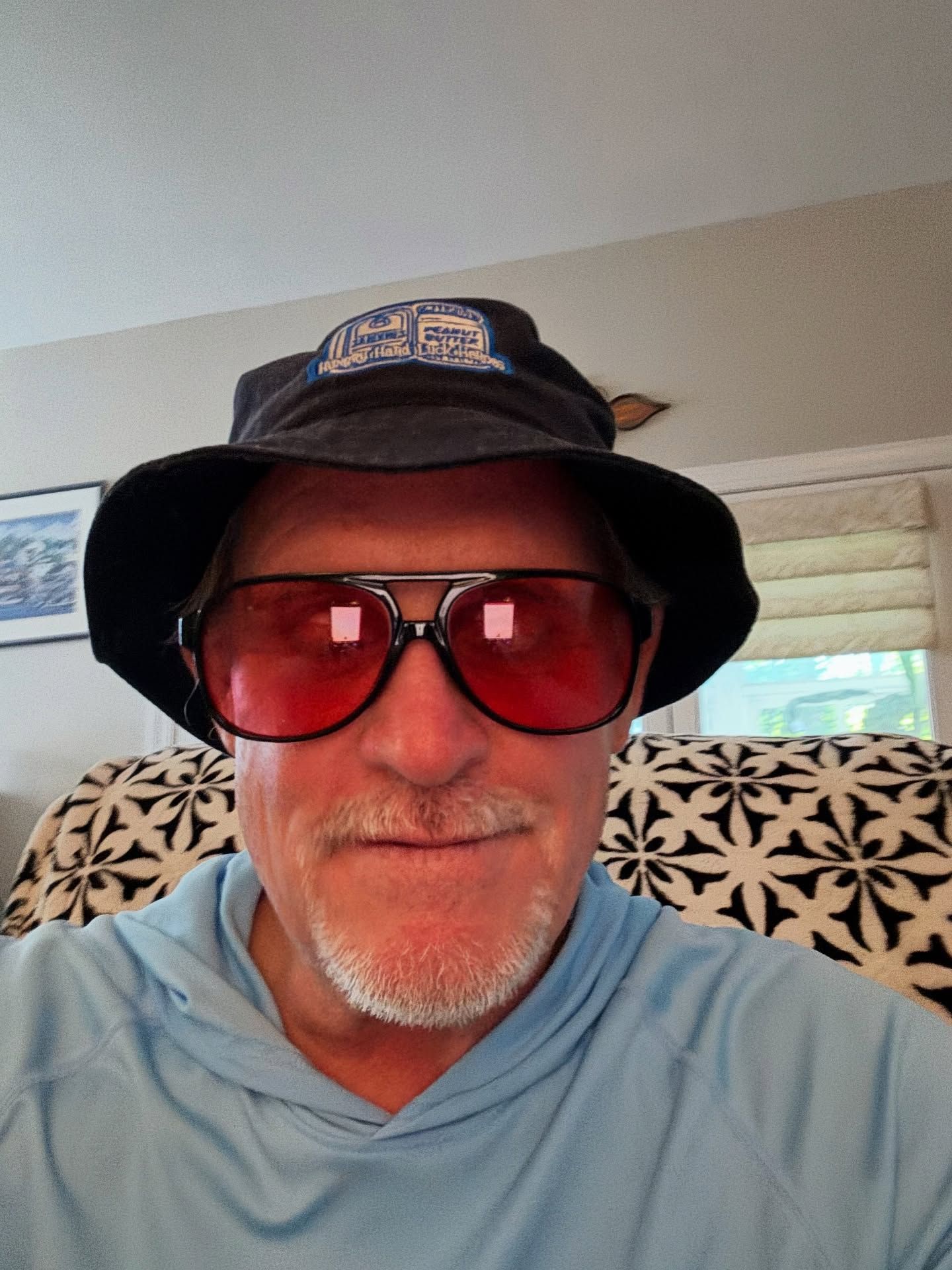 Man wearing hat and red-tinted aviator glasses, smiles slightly; indoors.