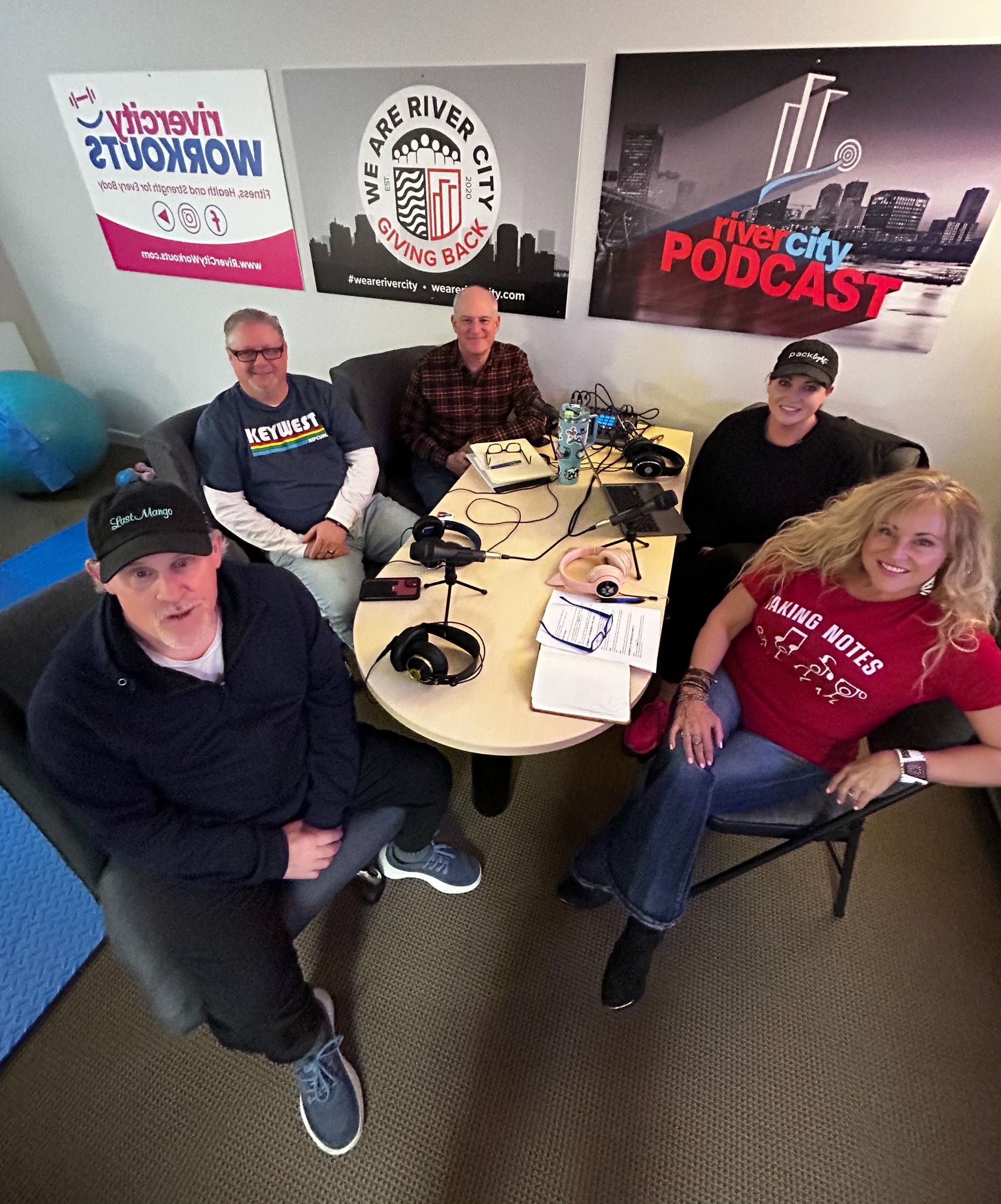 Group of people seated around a table, possibly for a podcast recording. Three posters in the background.
