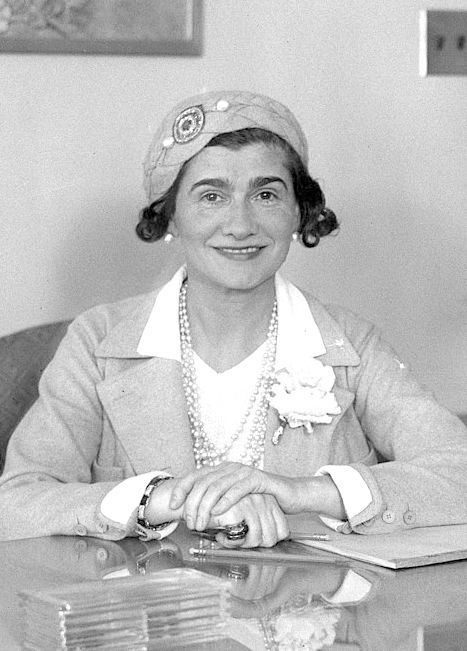 Woman in light-colored jacket and hat, seated at a desk, smiling with hands clasped.