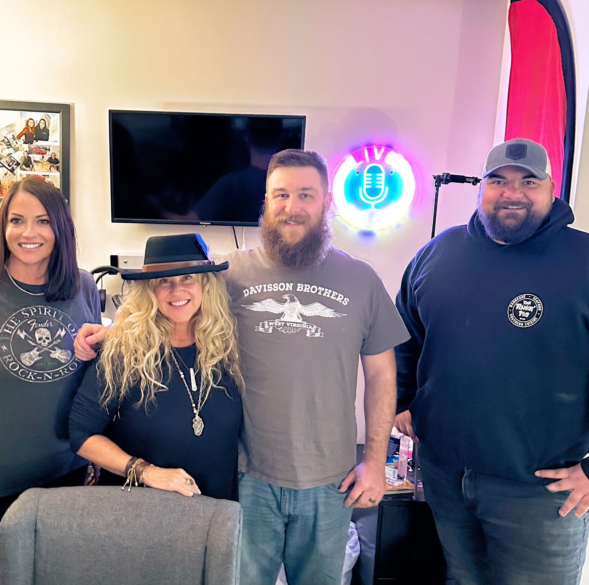 Four people smiling in front of a podcast setup with a TV, microphone, and neon sign.