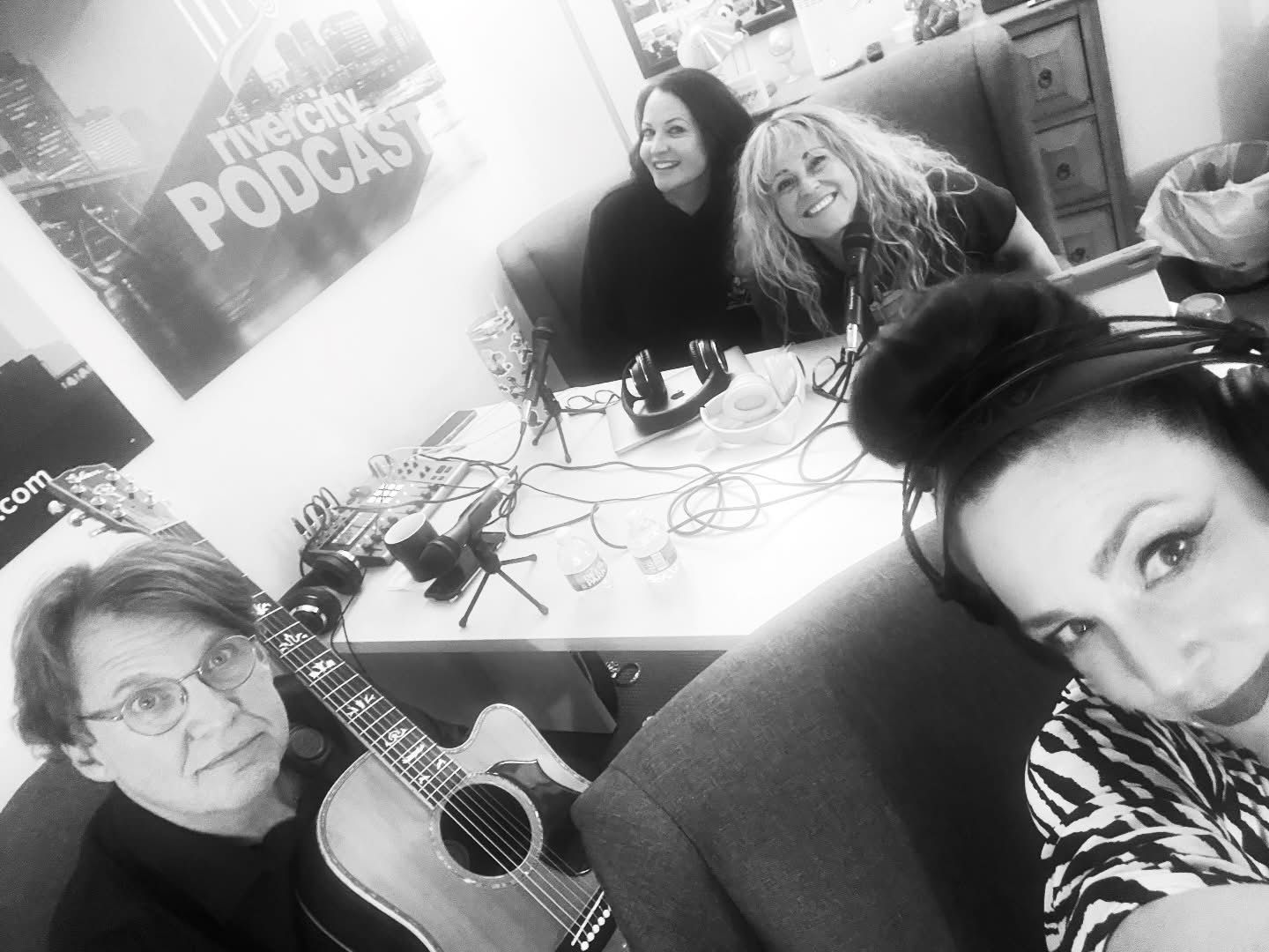 Four people in a room; one with a guitar, three near a table with audio equipment. All are smiling.