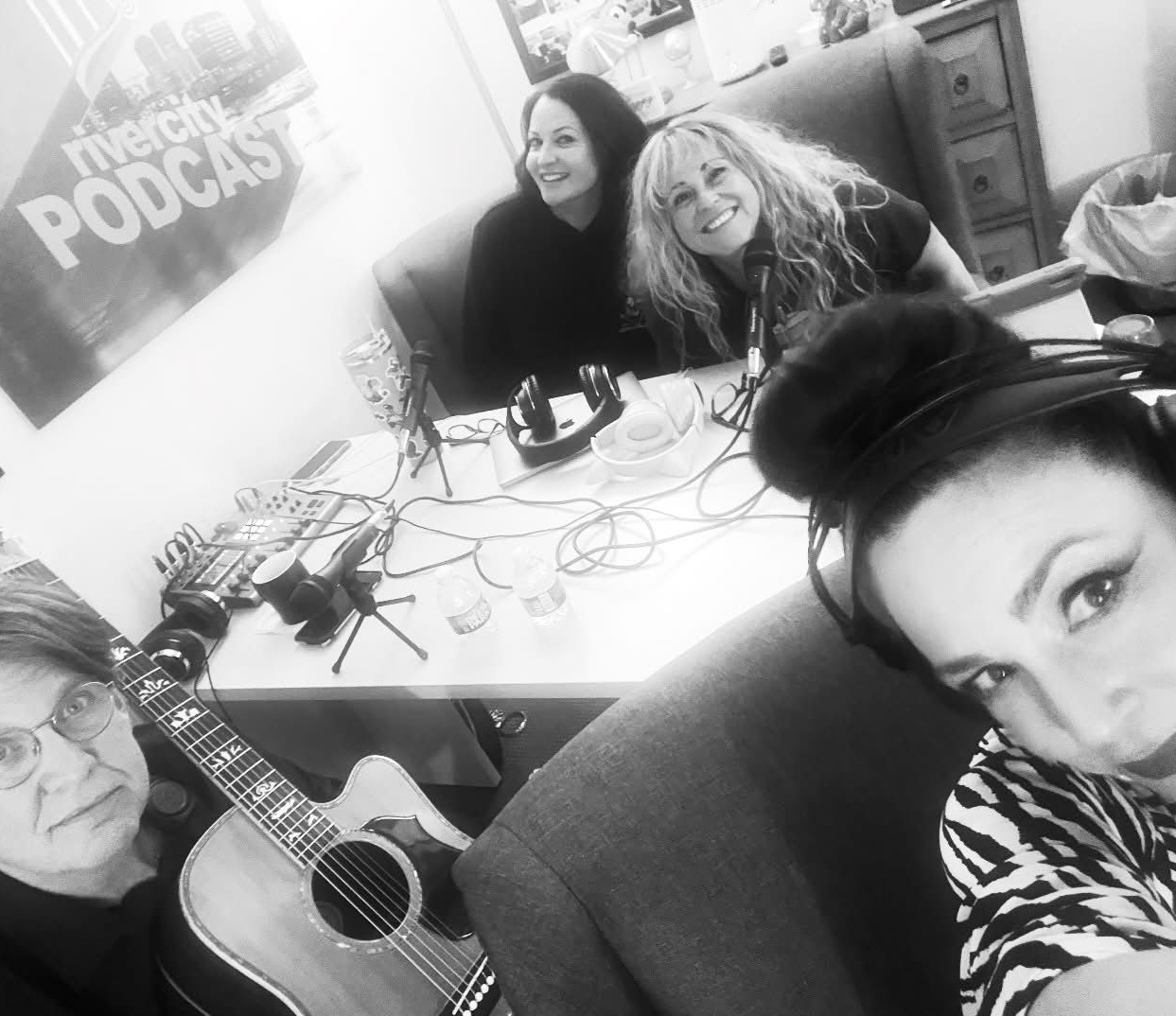 Four people in a room; two women smile at the camera. A man plays guitar. The fourth wears headphones. Podcast setup.