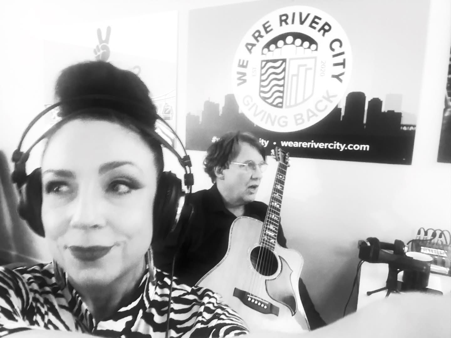 Woman with headphones and man with a guitar in front of a River City banner, both indoors.