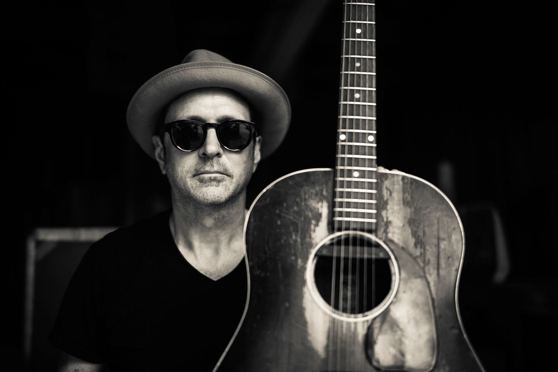A person wearing a fedora and sunglasses poses next to a well-worn acoustic guitar in a dimly lit, high-contrast setting.