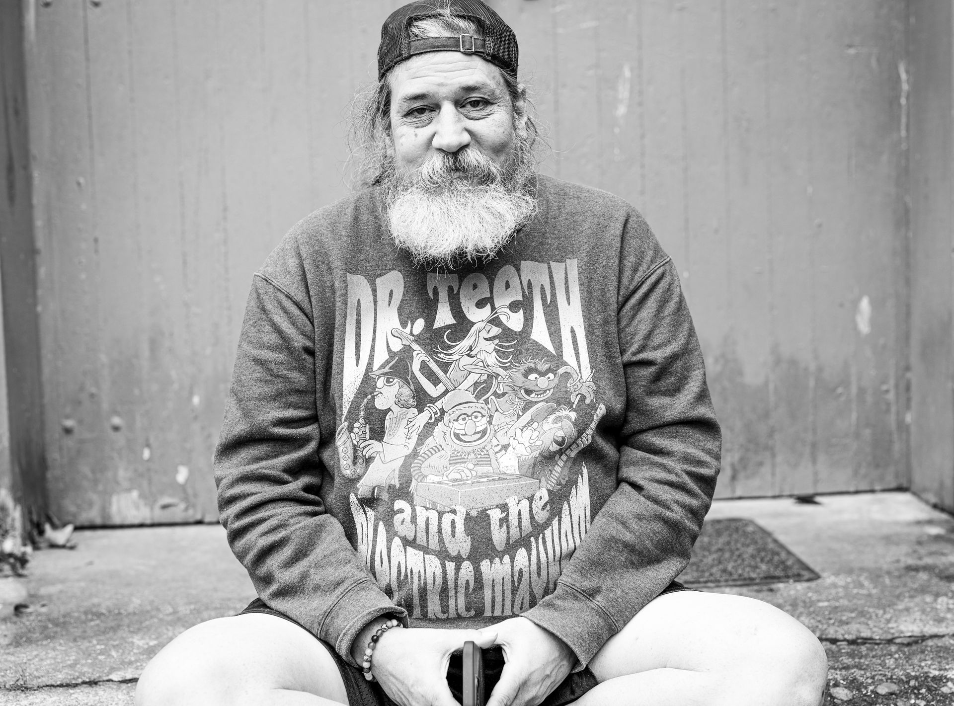A person with a beard, wearing a baseball cap and a Dr. Teeth and the Electric Mayhem sweatshirt, sits cross-legged.