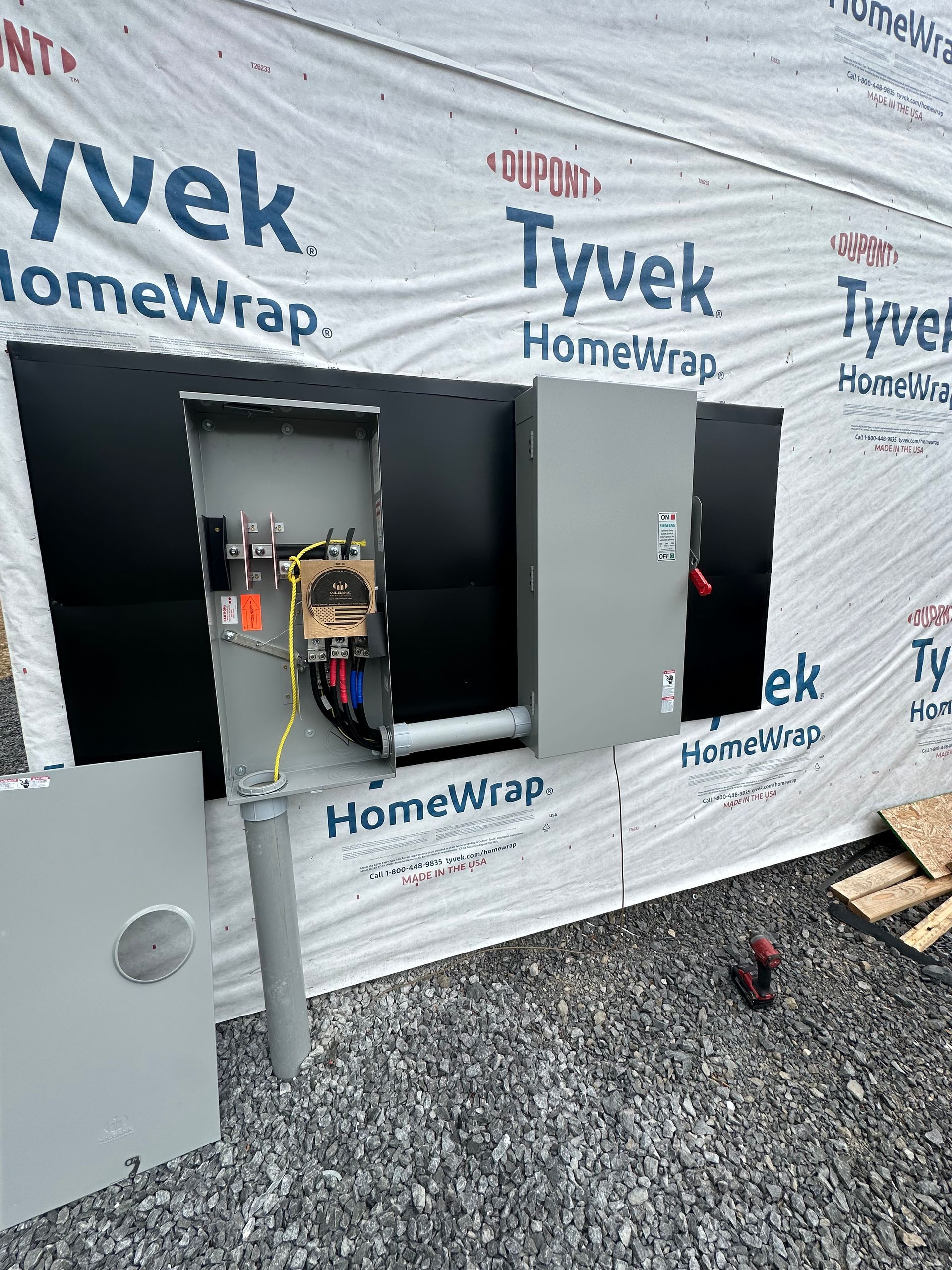 A electrical box is sitting on the ground in front of a building covered in tyvek homewrap.