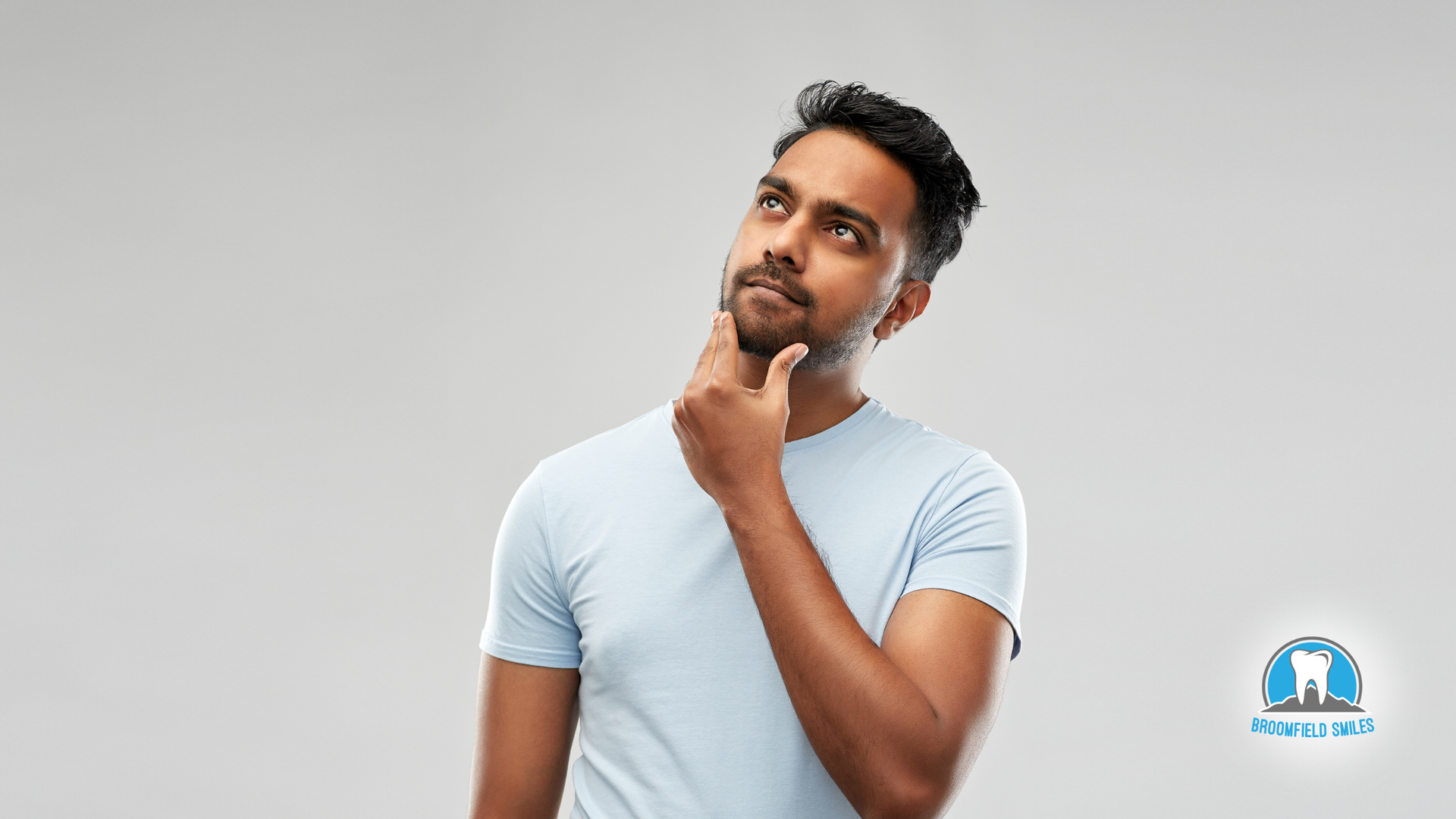 A man in a white t-shirt is holding his hand to his chin and looking up.