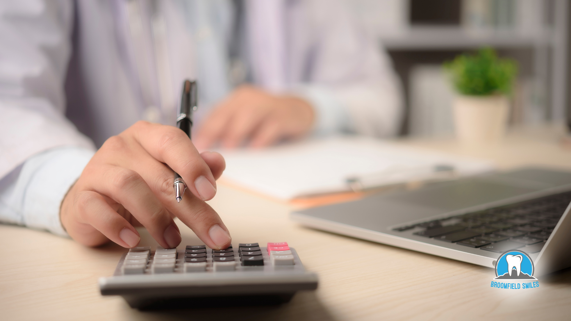 Person in lab coat using a calculator at a desk with a laptop and paperwork.