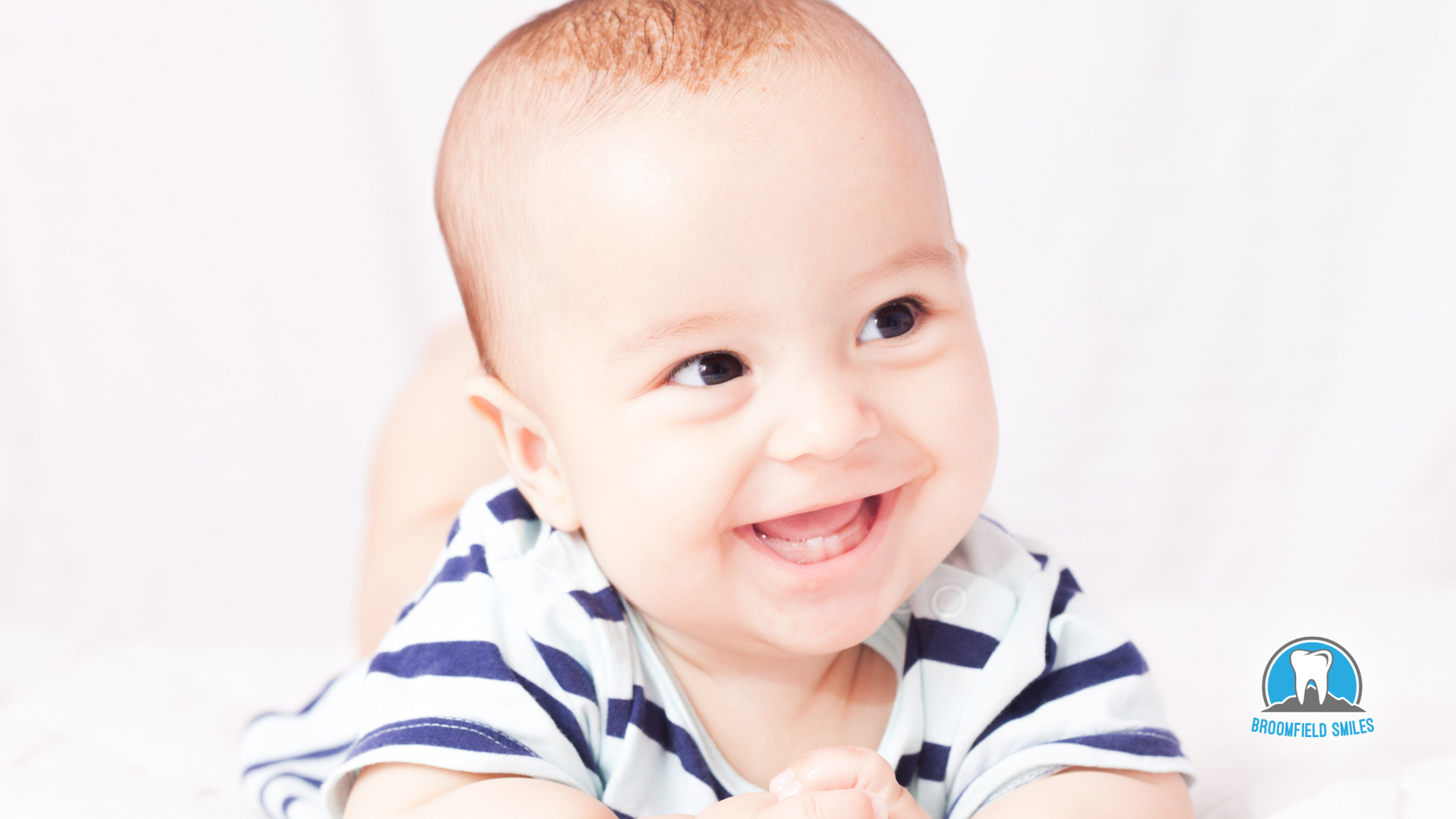 A baby is smiling while laying on his stomach on a bed.
