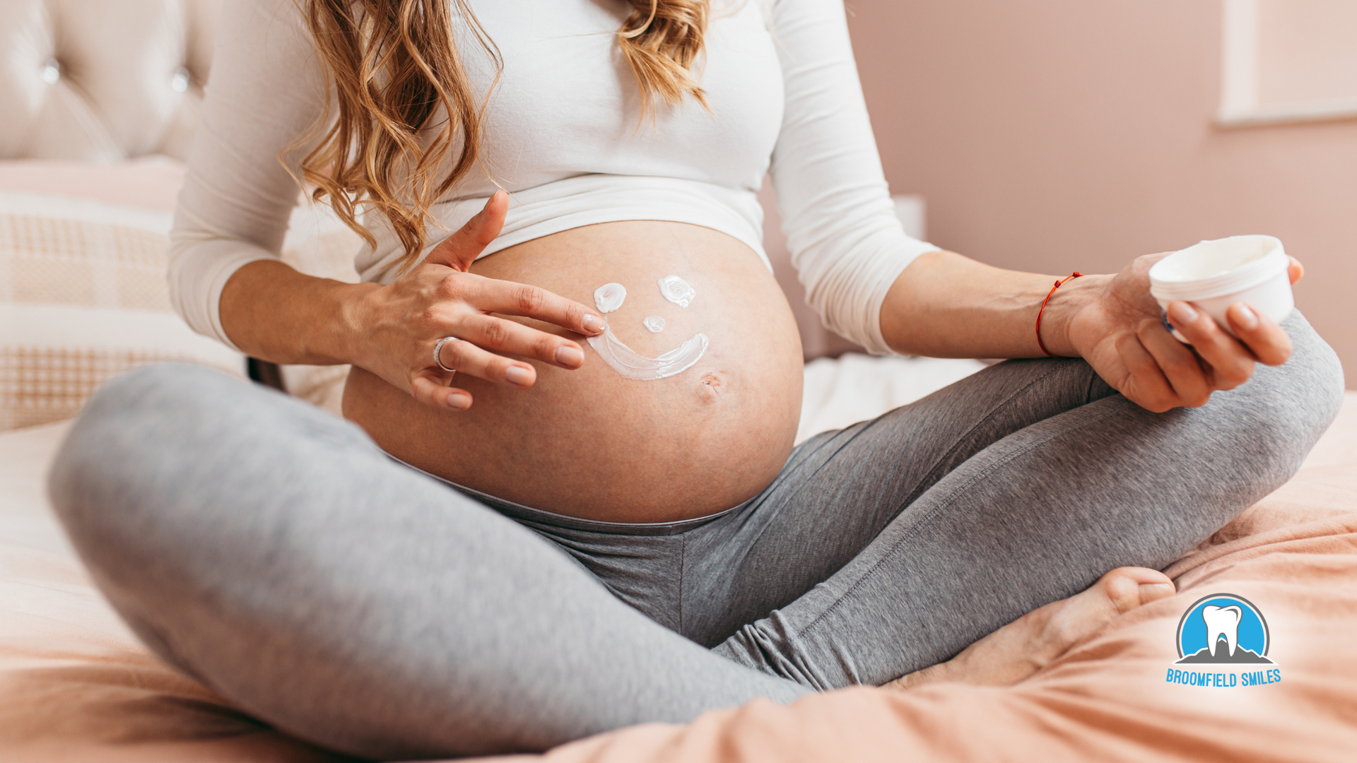 A pregnant woman is sitting on a bed applying lotion to her belly.