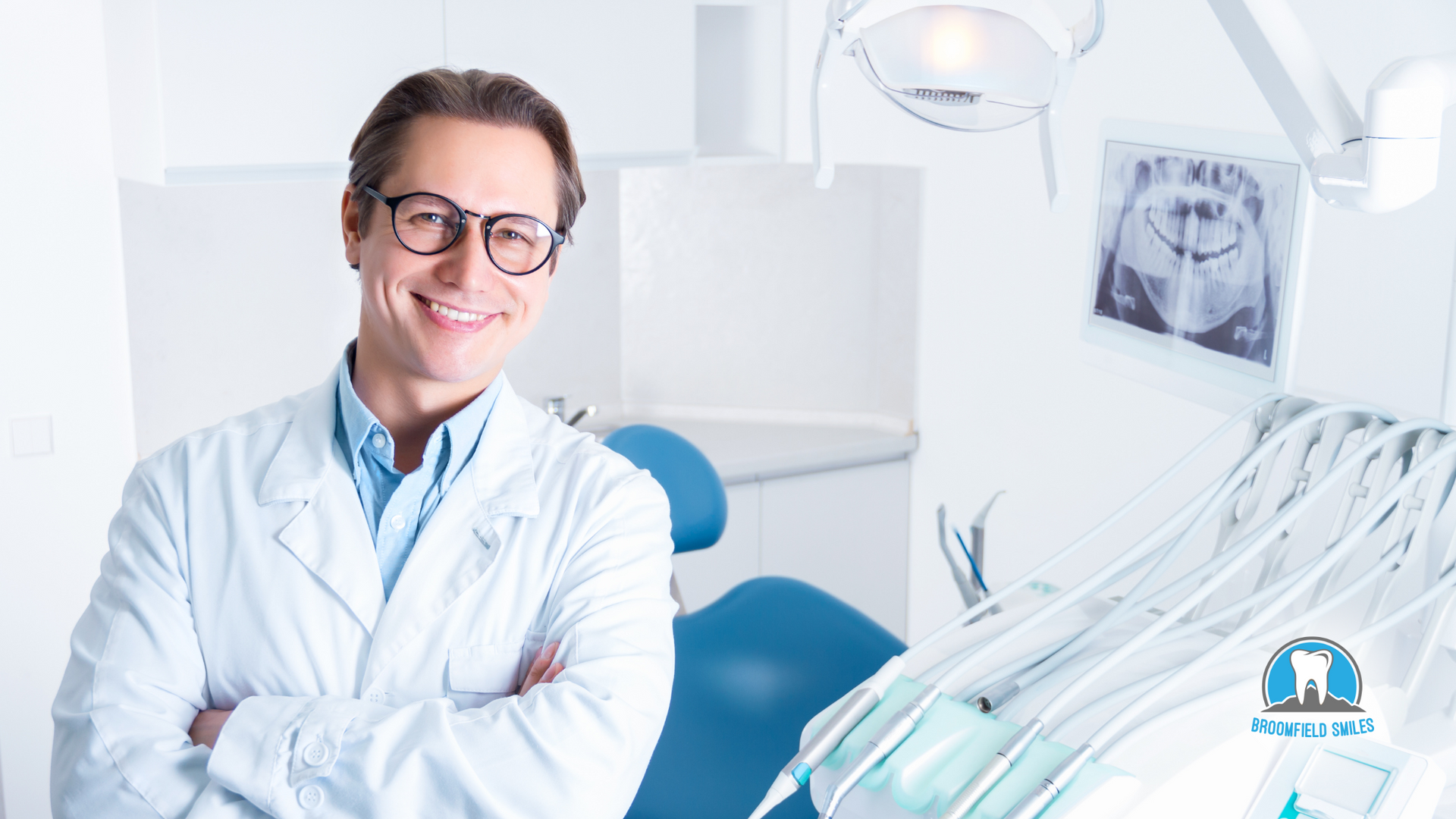 A dentist is smiling in a dental office with his arms crossed.