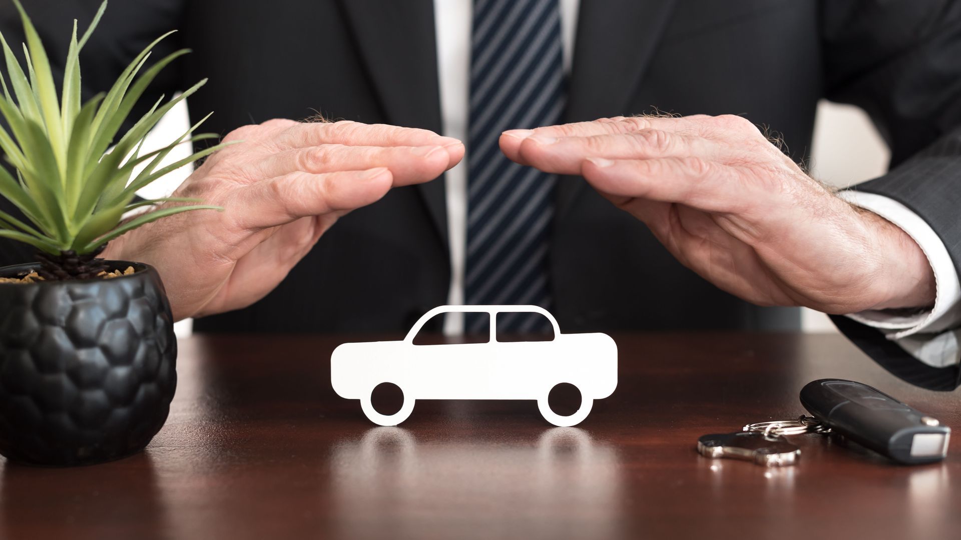 Hands of a person in a suit protecting a small car cutout on a table, with car keys and a potted plant.