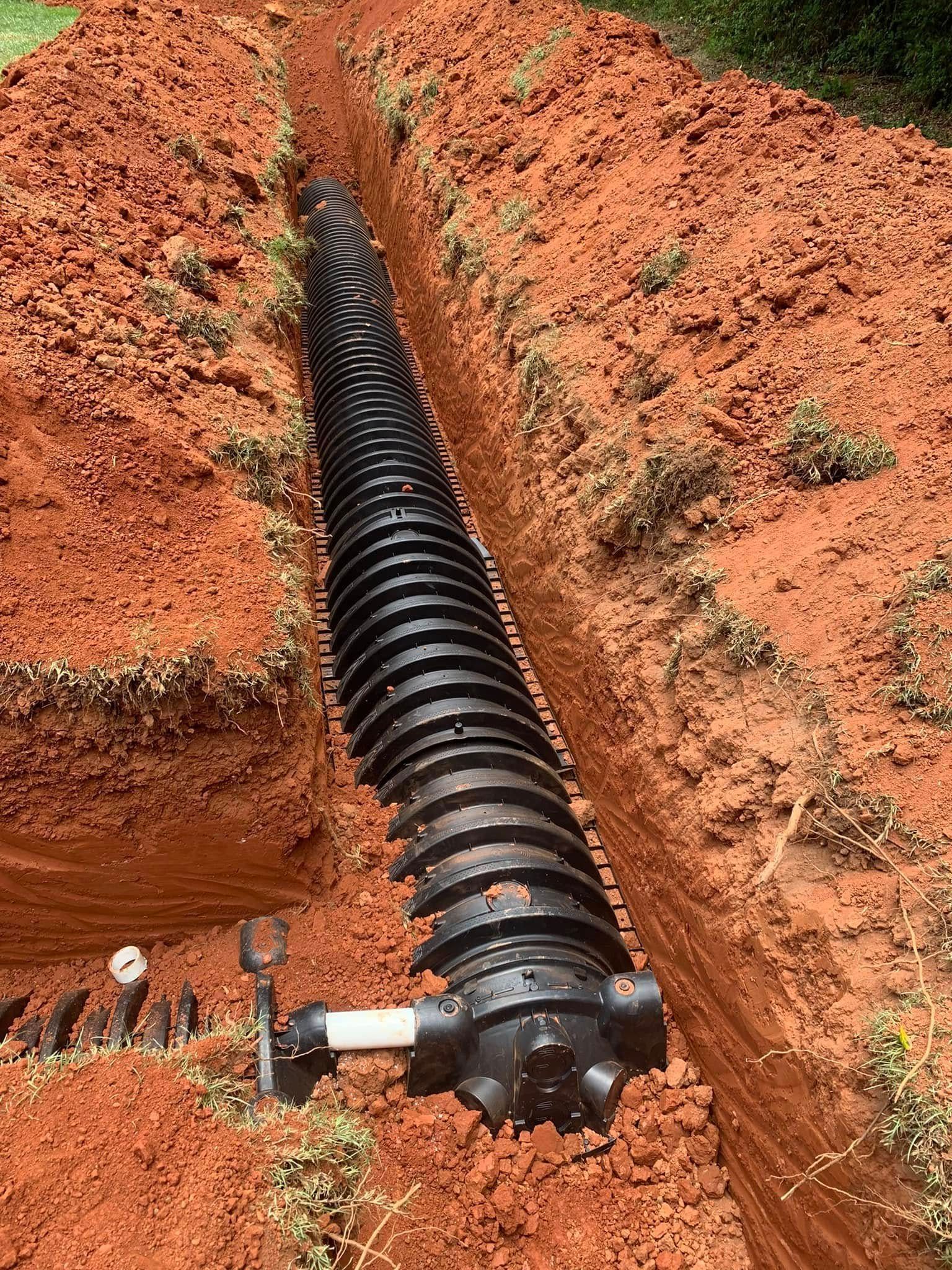 Black corrugated drain pipe in a trench, surrounded by red soil, with a white pipe connection visible.