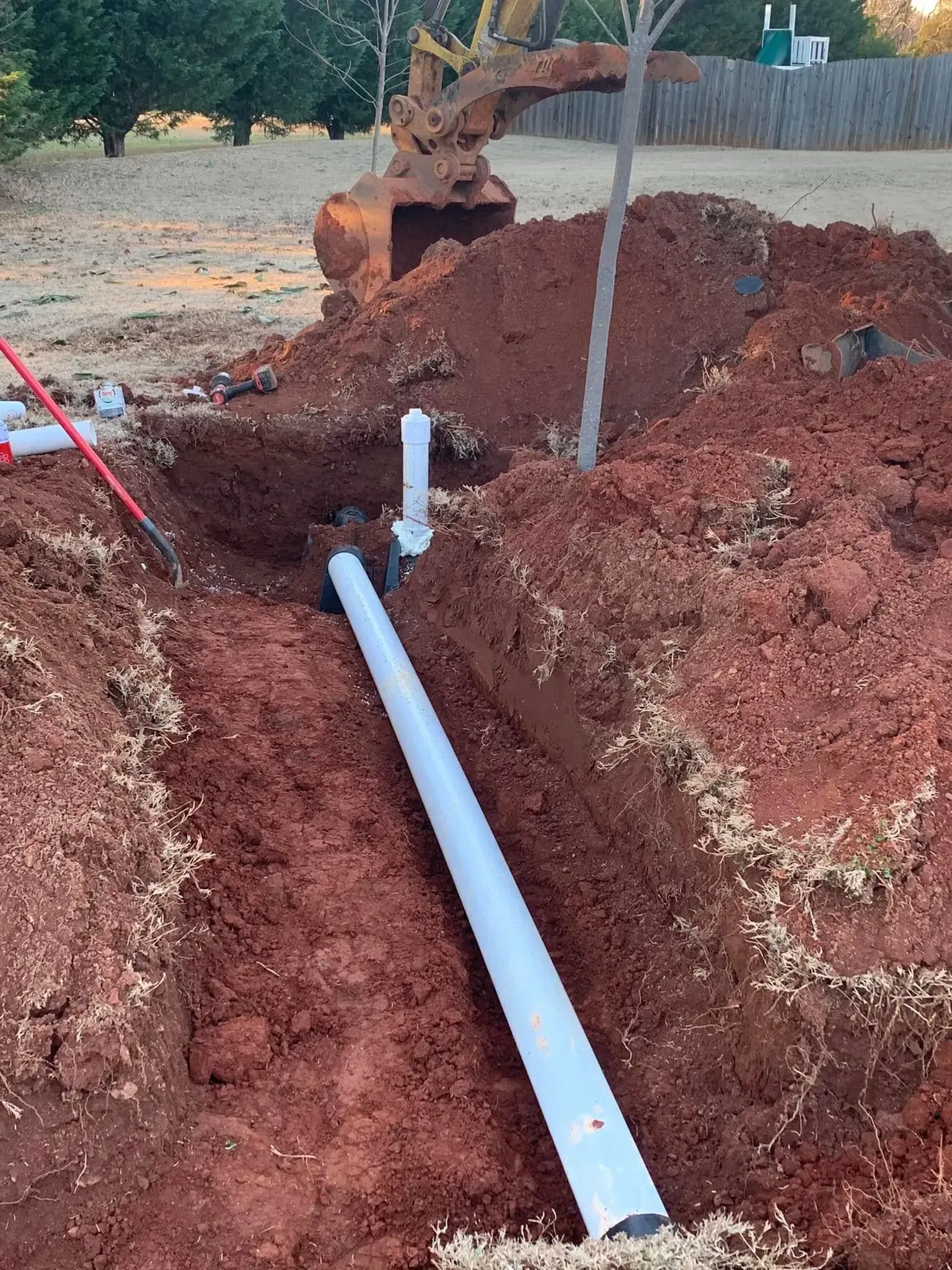 Trench dug in red soil, with PVC pipe and backhoe preparing to install plumbing.