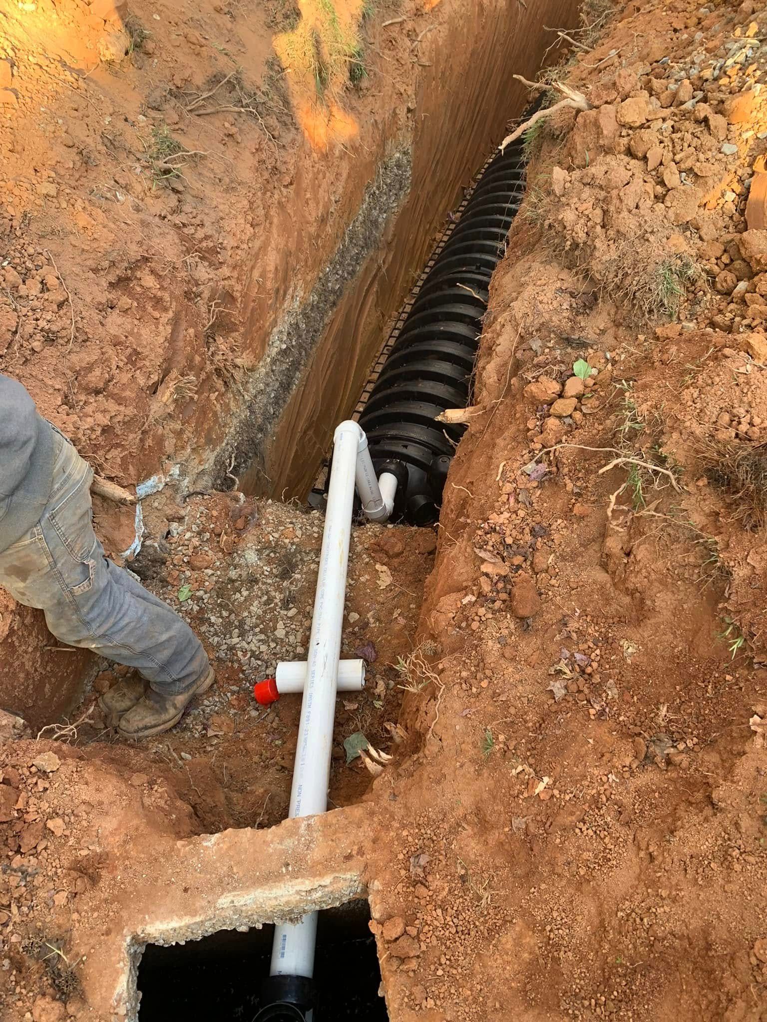Trench with black corrugated pipe and white PVC plumbing being installed by a person in a dirt setting.