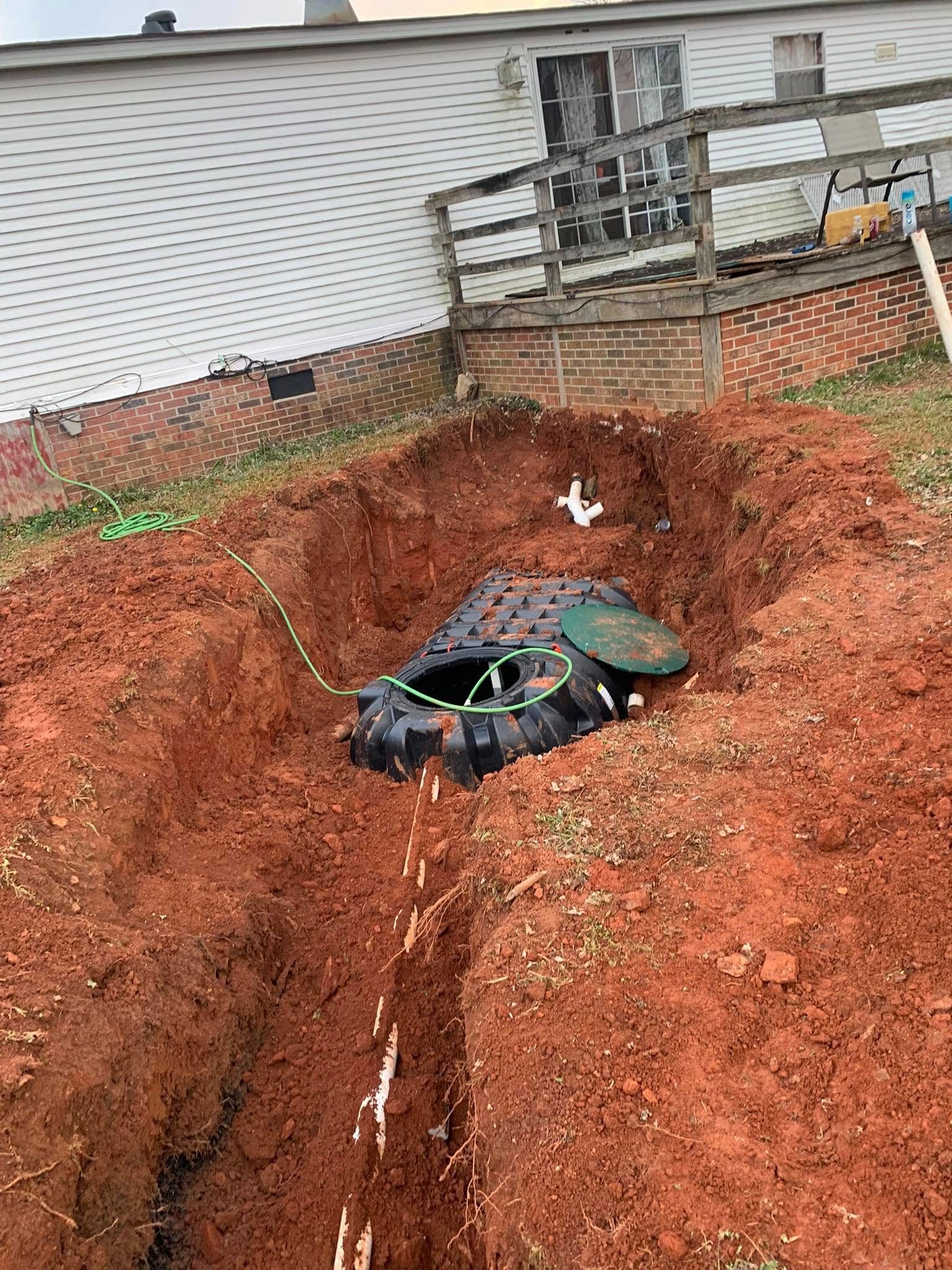 Septic tank in an excavated hole. Red soil surrounds a partially open black tank with green tubing.