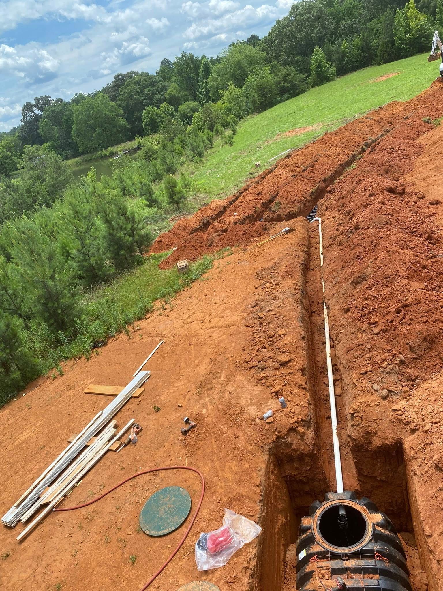 Trench dug in red soil, with white pipes and a black tank visible. Green vegetation and blue sky in background.