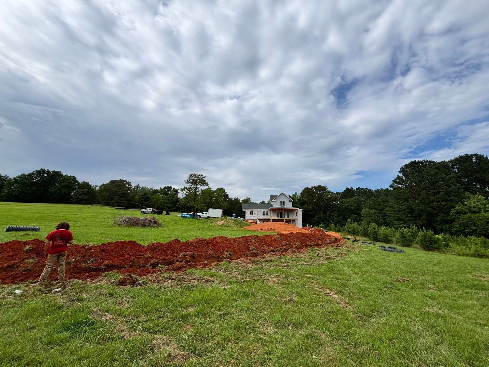 Person working on construction site; house in the distance, red soil, green grass, cloudy sky.