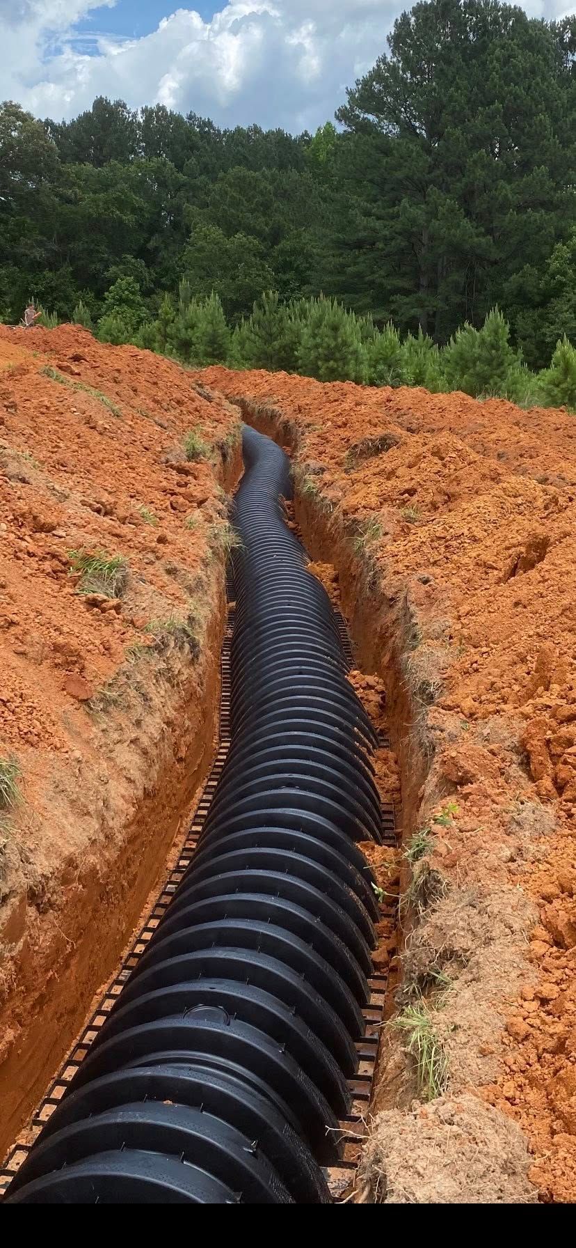 Black corrugated pipe in a trench of red dirt, likely for drainage, with trees and sky in the background.