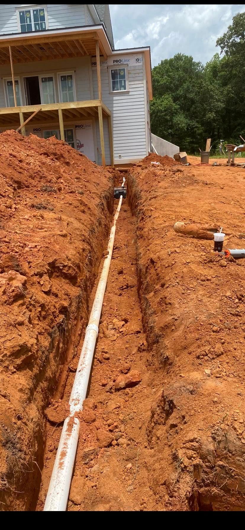 A white pipe in a trench runs towards a house under construction in red soil.