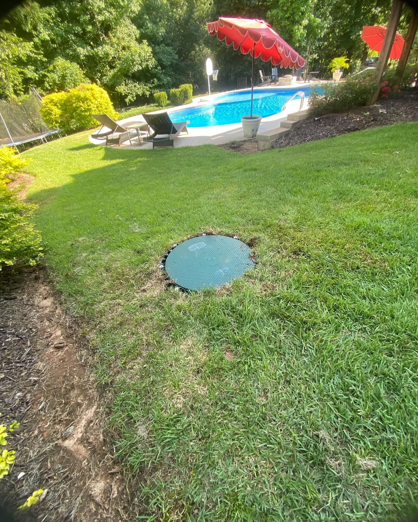 Green circular septic tank cover in a grassy yard, with a pool and umbrella in the background.