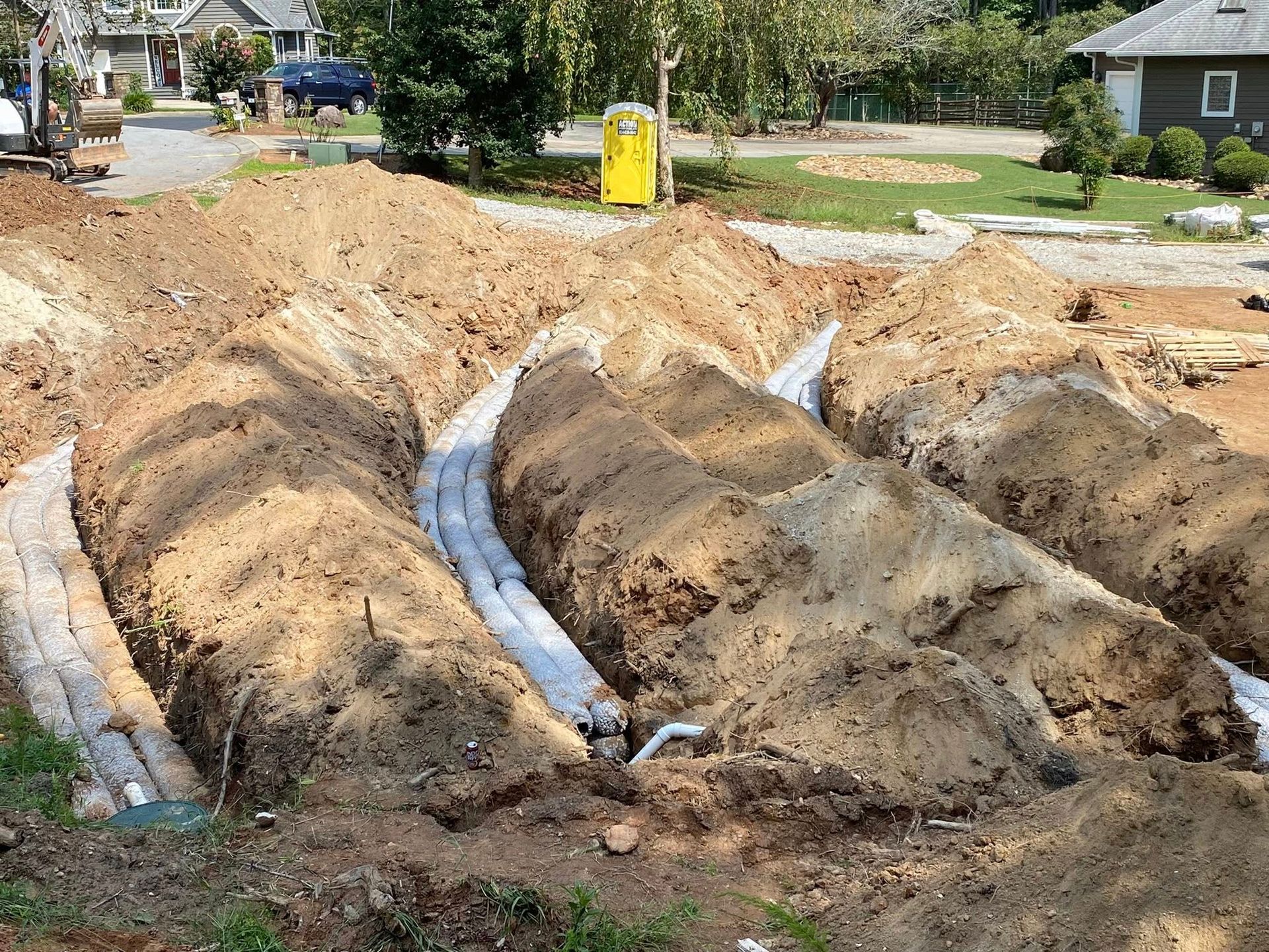 Trenches dug in brown earth with white pipes visible, likely for underground utility installation, near a house.