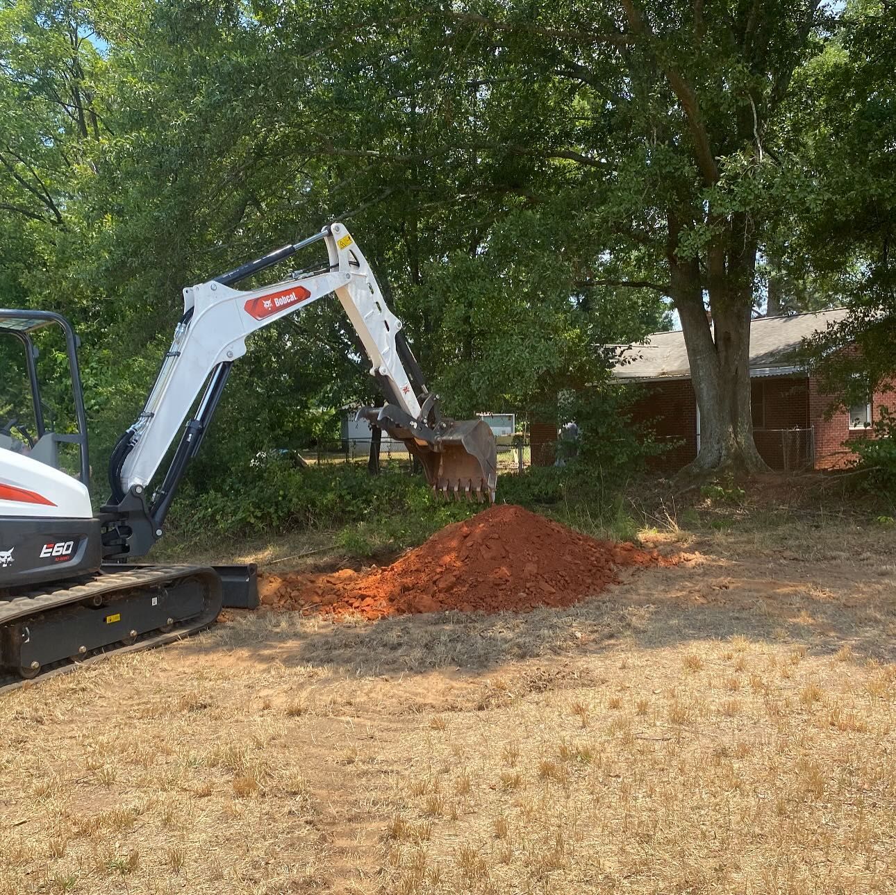 White Bobcat excavator digging red dirt in a yard near a house and trees.