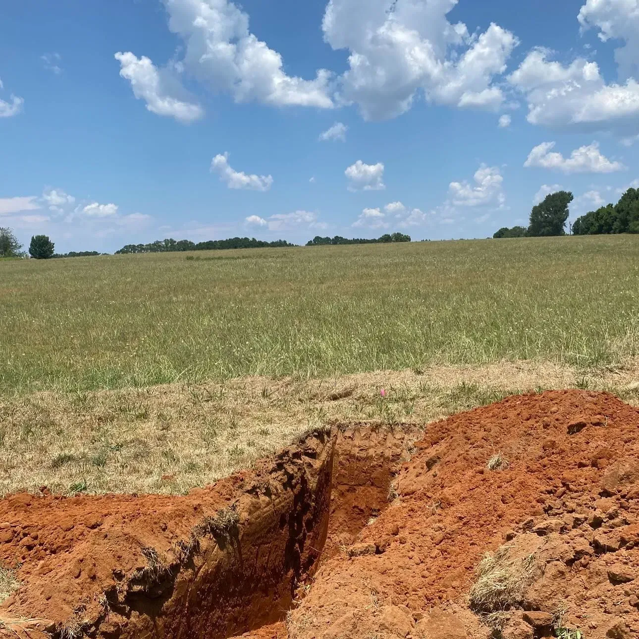 A dug trench in a field with a blue sky and scattered clouds overhead.