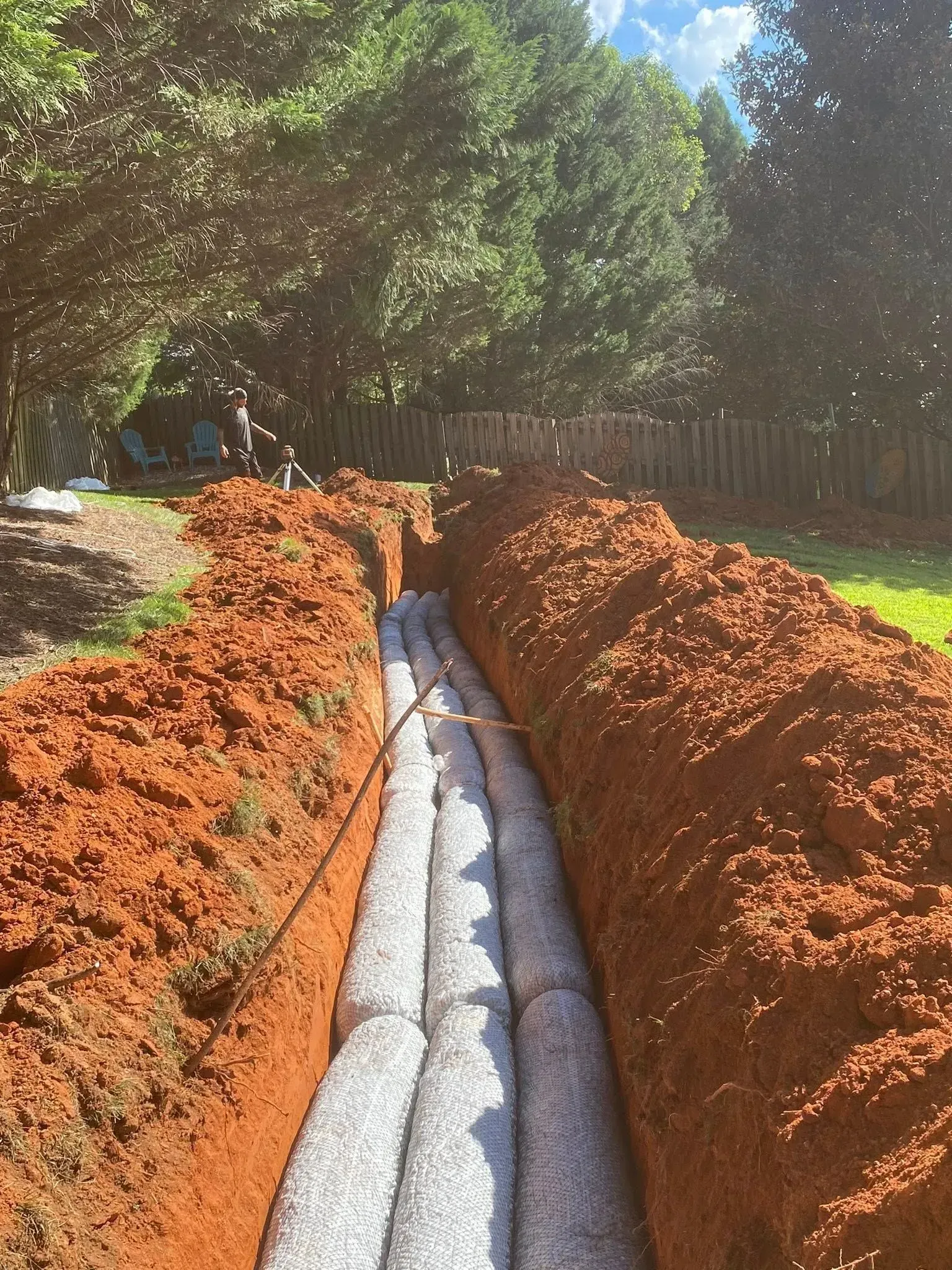 Trench filled with grey gravel-filled tubes, likely for drainage, in red soil, outdoors.