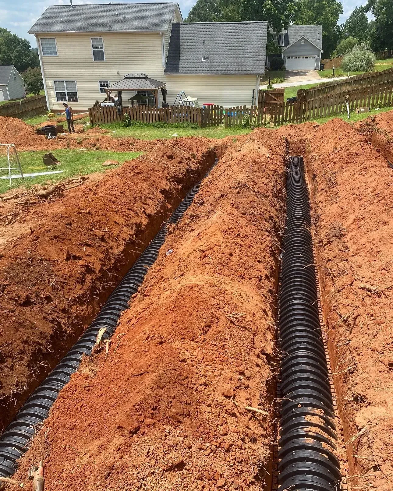 Two parallel trenches with black corrugated pipes installed, prepared for underground drainage in a residential yard.