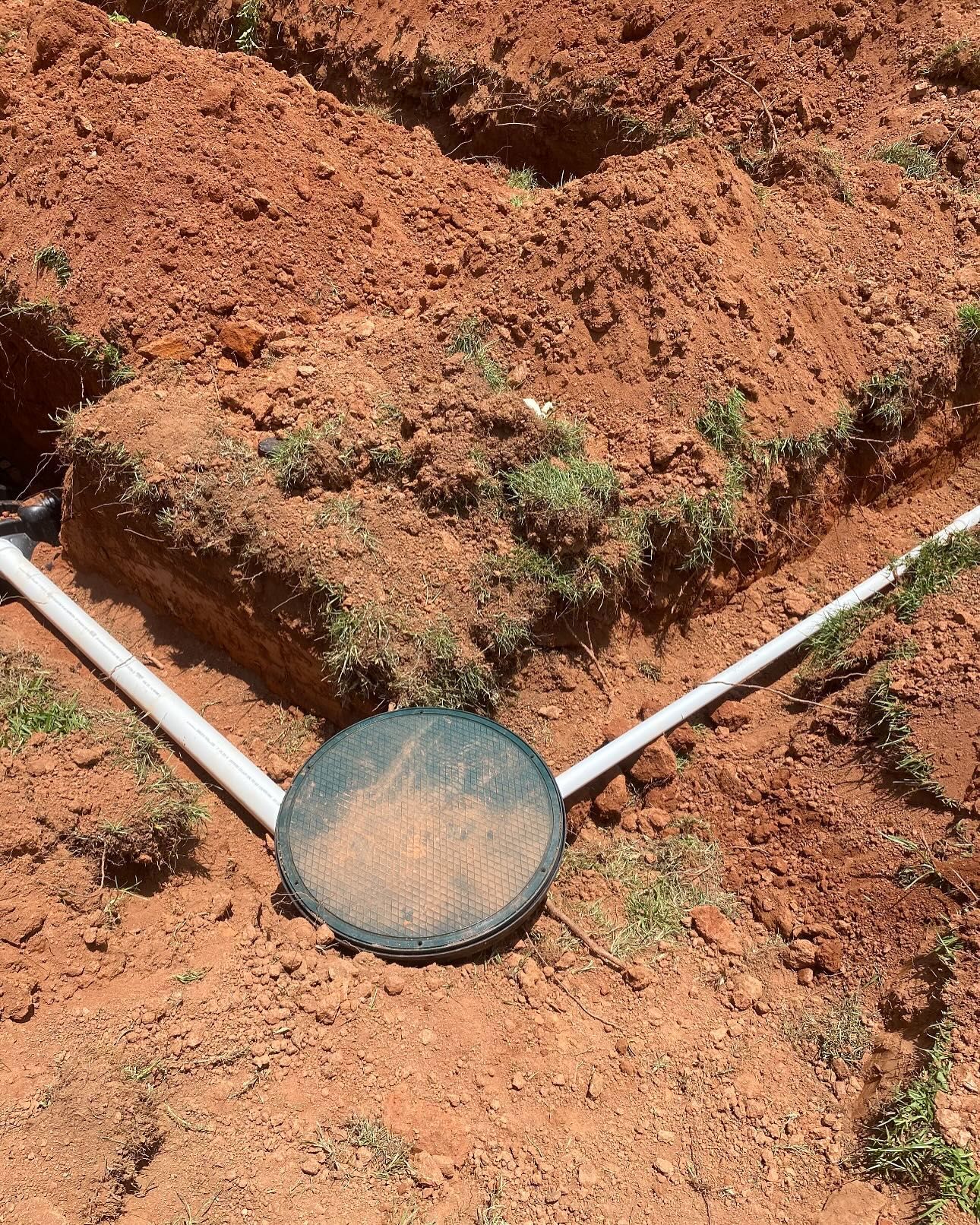 Trenches dug in red dirt with a white PVC pipe system and a circular lid.