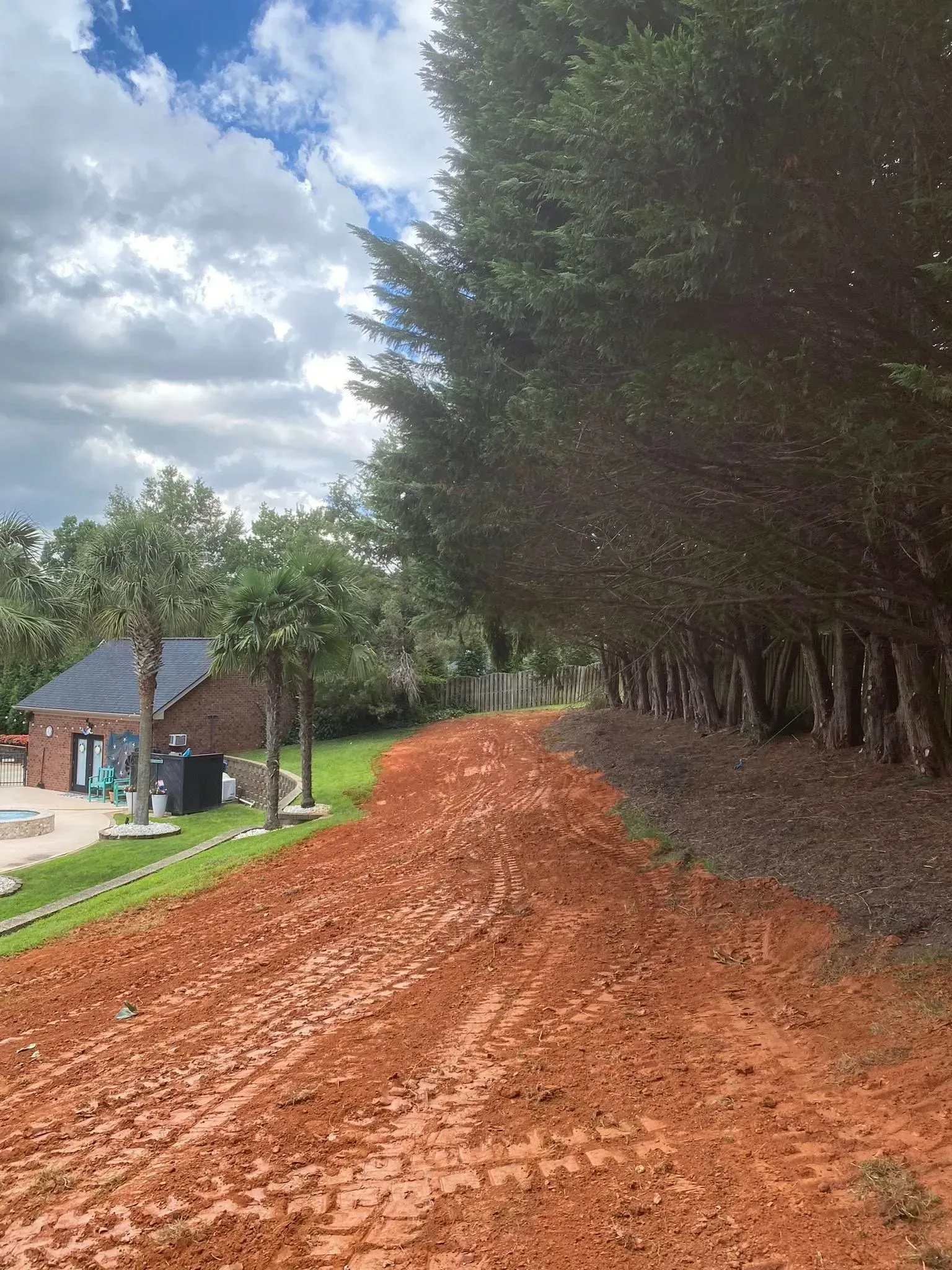 Muddy, red-brown dirt path next to a line of trees on a slight incline; a house and sky are in the background.