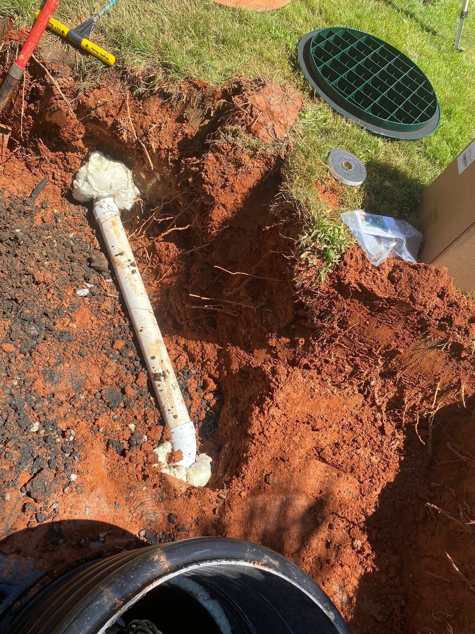 Hole in red dirt with a white pipe and concrete base, near a green cover and tools, outdoors.
