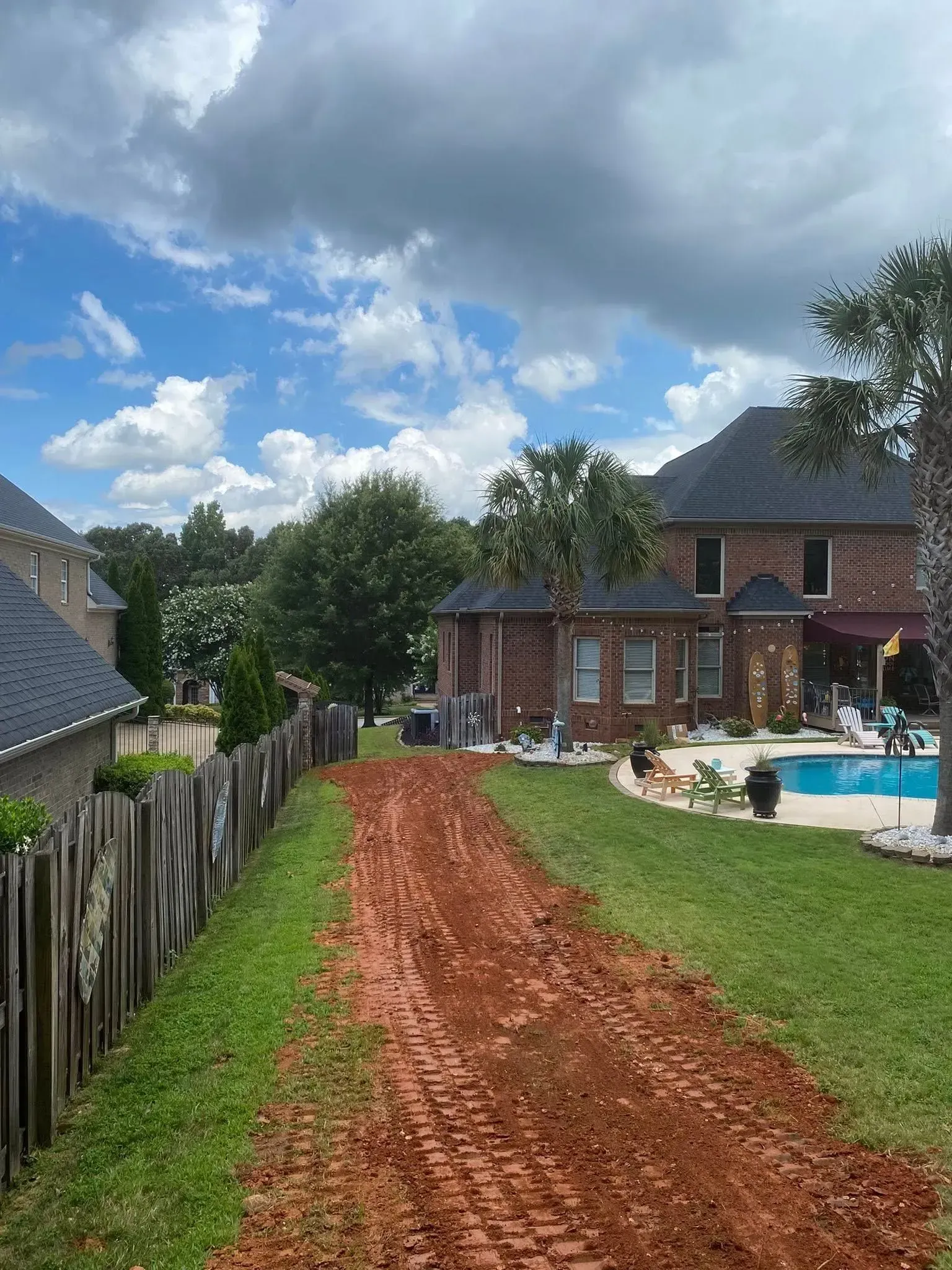 Dirt path through a grassy yard toward a house with a pool on a sunny day.
