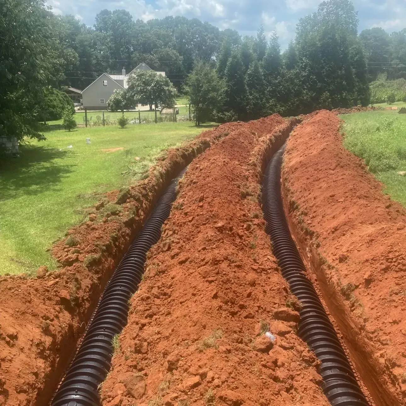 Two black corrugated pipes in parallel trenches dug in red soil, with a grassy lawn and trees in the background.