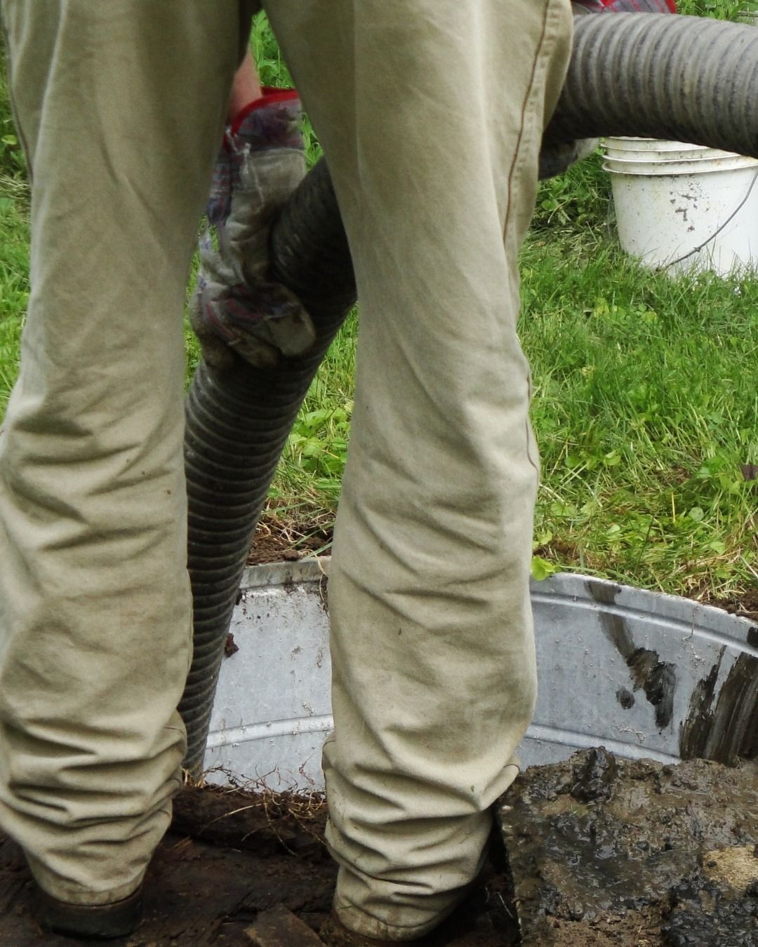 Person in tan pants holds a hose over a septic tank, near grass and a white bucket.